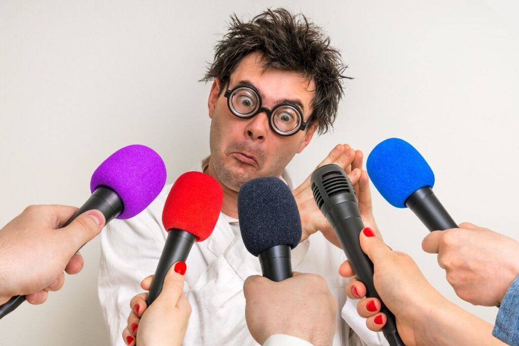 A stereotypical mad scientist in a white lab coat surrounded by media microphones